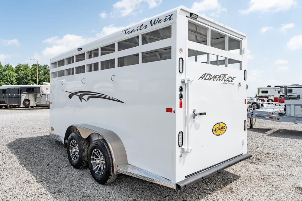 Back and side view of a white Trails West Adventure horse trailer with dual wheels and decorative graphics, parked on a gravel lot.