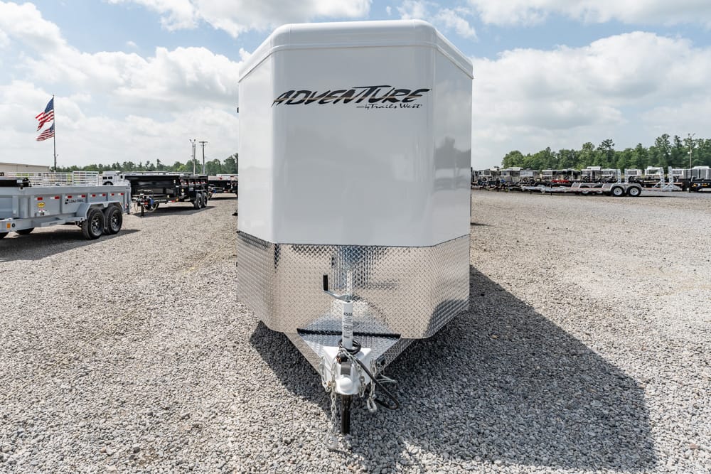 Front view of a white enclosed cargo trailer with diamond-plate lower front and hitch, labeled Adventure, on a gravel lot.