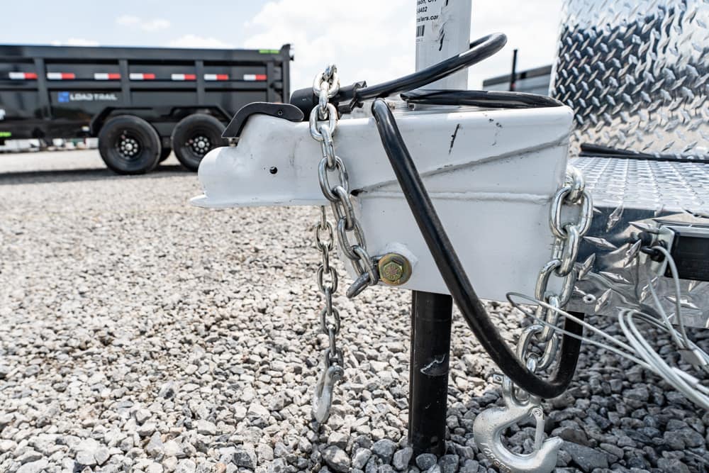 Close-up of a trailer hitch with chains and a metal jack on a gravel lot; a dump trailer is visible in the background.