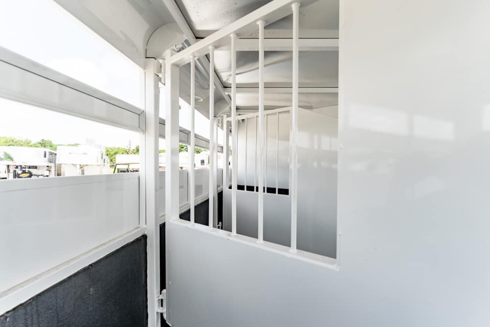 Interior of a white horse trailer with vertical metal bars forming stalls and a gray floor, viewed from between the partitions.