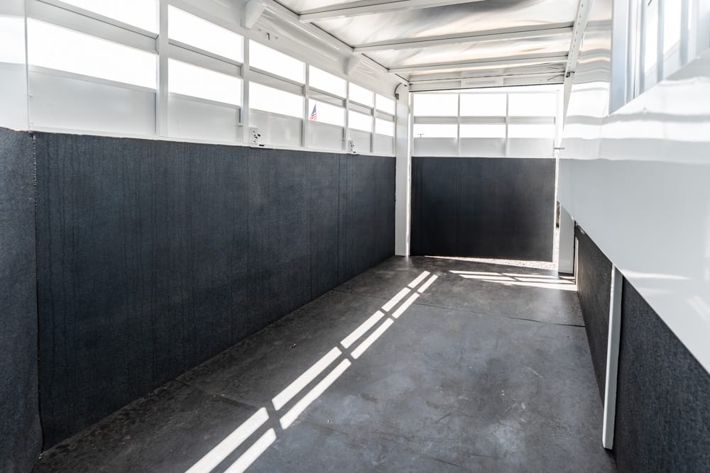 Interior of an empty white horse trailer with dark padded walls and sunlight streaming through windows in the ceiling and sides.