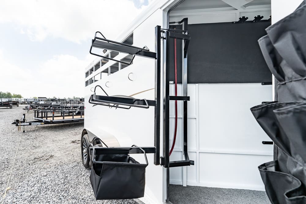 Open horse trailer with fold-out saddle racks and gear bags, parked on a gravel lot.