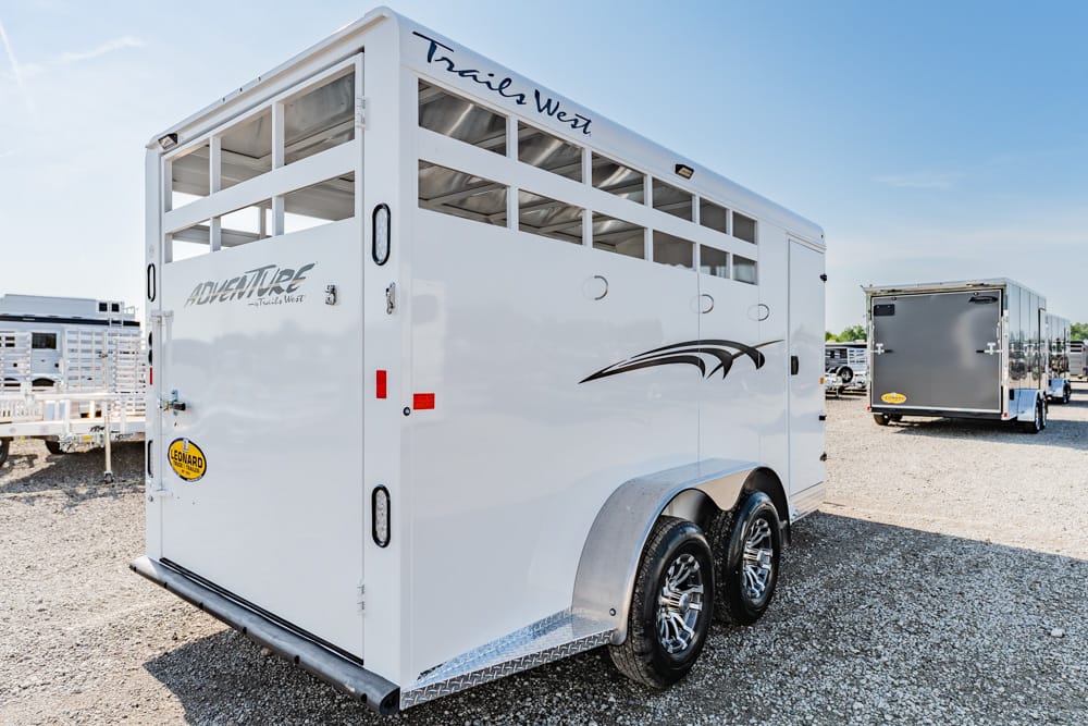 White enclosed horse trailer with two axles and chrome wheels, parked on a gravel lot under a clear sky, labeled Adventure by Trails West