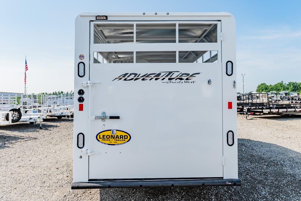 Rear of a white trailer with an 'ADVENTURE' logo, Leonard Trailer sticker, and a gravel lot in the background.
