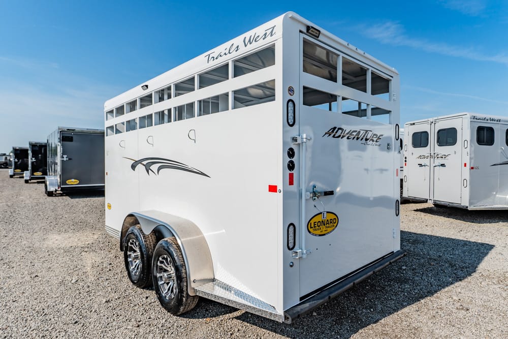 White dual-axle horse trailer with windows along the top and the word 'Adventure' on the back door, parked on gravel under a blue sky.