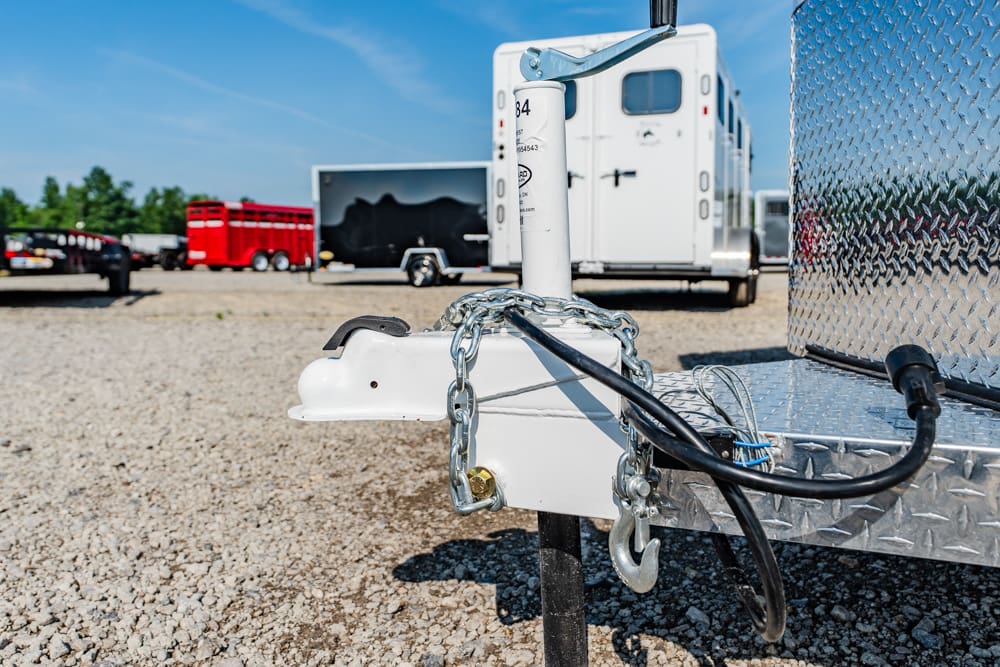 Close-up of a trailer hitch with safety chains and electrical cable on a gravel lot; white trailer tongue with background trailers.