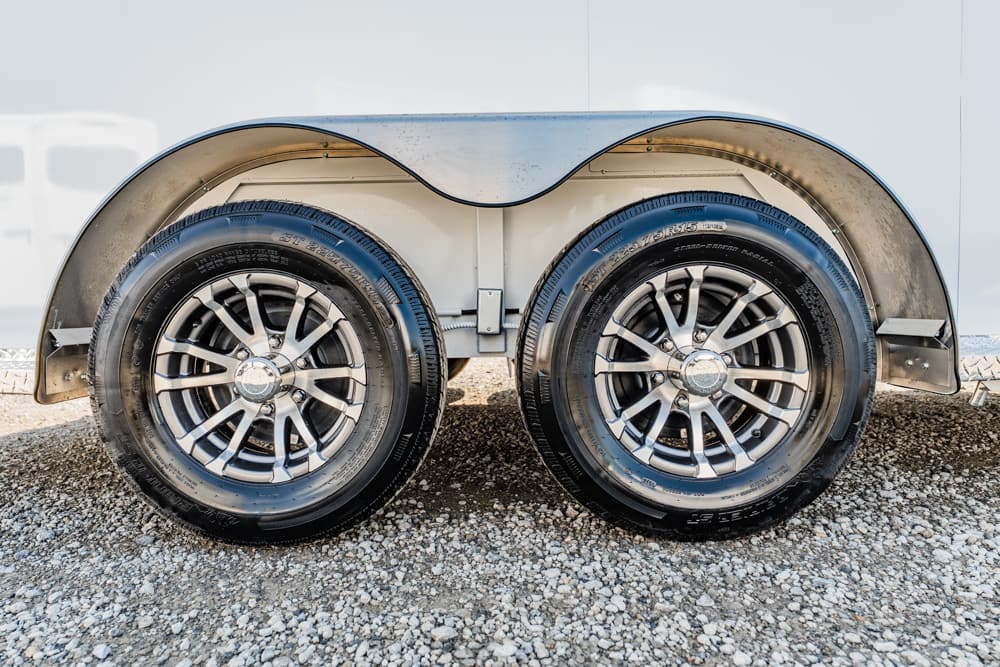 Double-axle trailer wheels with chrome rims resting on a gravel surface under a silver fender mudguard