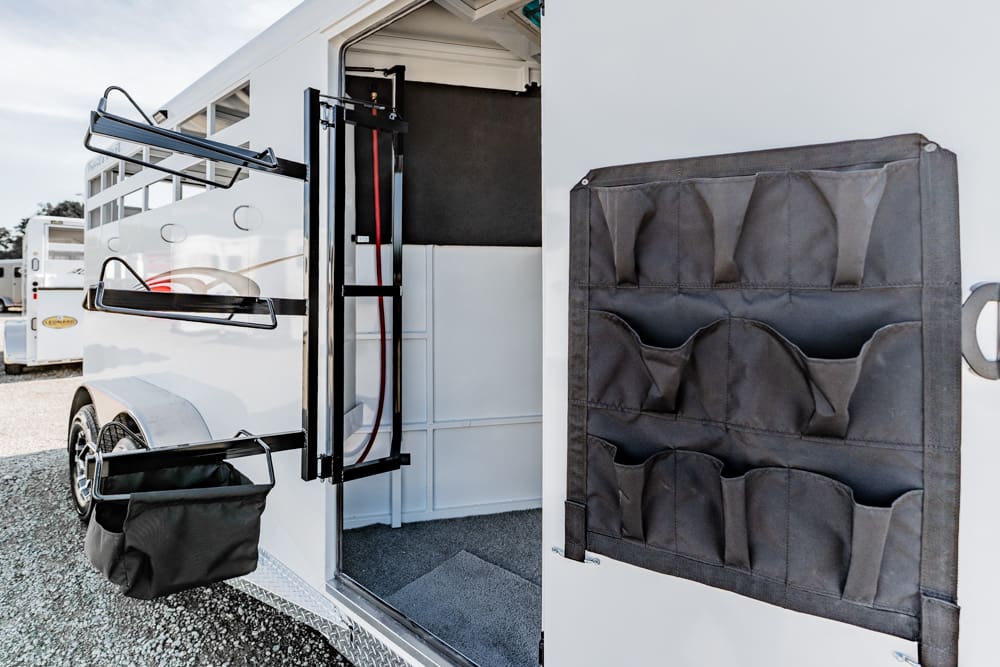 Open white horse trailer with a door-mounted saddle rack and hanging storage pockets on the interior wall of the doorway.