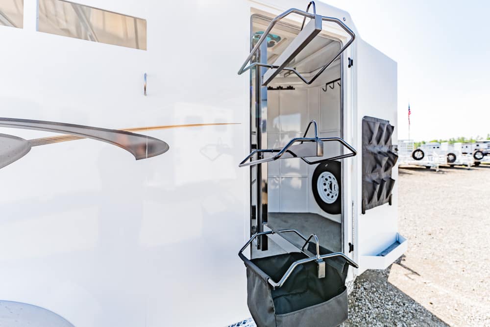 Open rear of a white enclosed trailer with hanging metal racks on the door and a gray storage bag at the bottom; spare tire visible inside.