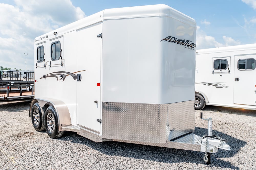 White horse trailer with diamond-plate front and two axles, labeled 'Adventure,' parked on gravel lot.