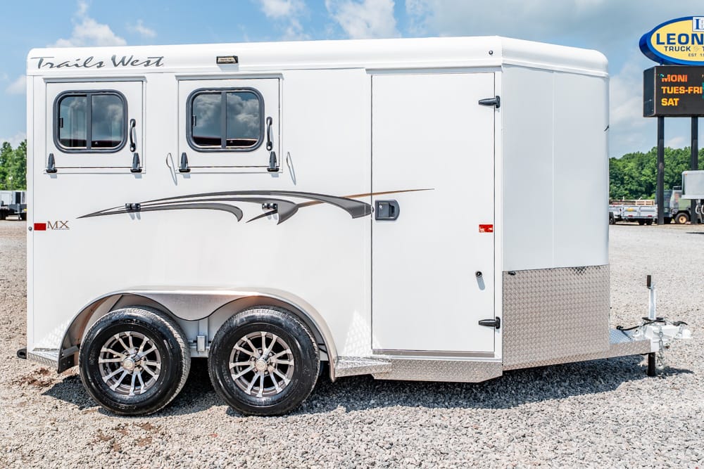 White enclosed horse trailer with two windows, decorative graphics, dual axles, and a front hitch on a gravel lot