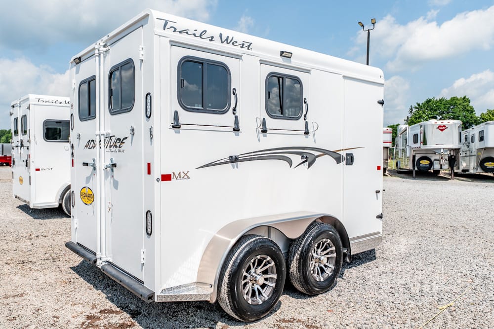 White horse trailer with dual axles and decorative graphics parked on a gravel lot, Trails West logo visible on the top edge of the trailerchained in the background.