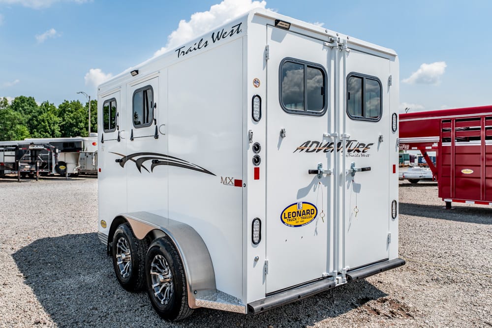 Rear view of a white Trails West Adventure horse trailer with dual doors, two small windows, and chrome wheels on a gravel lot.