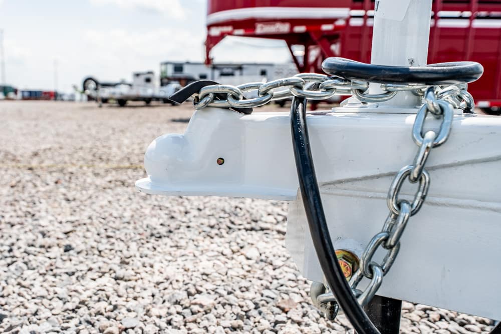 Close-up of a white trailer hitch secured with chain and a lock on a gravel lot, with a red truck in the background.