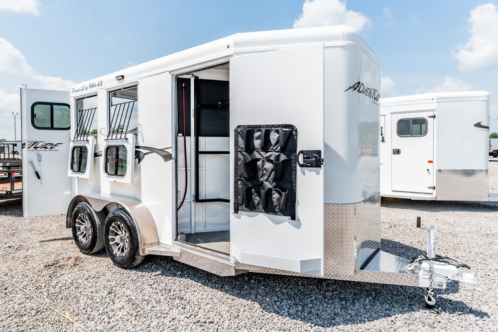 White tandem horse trailer with open side access, showing windows, stalls, and a storage pouch on the front, parked on gravel under a blue sky.