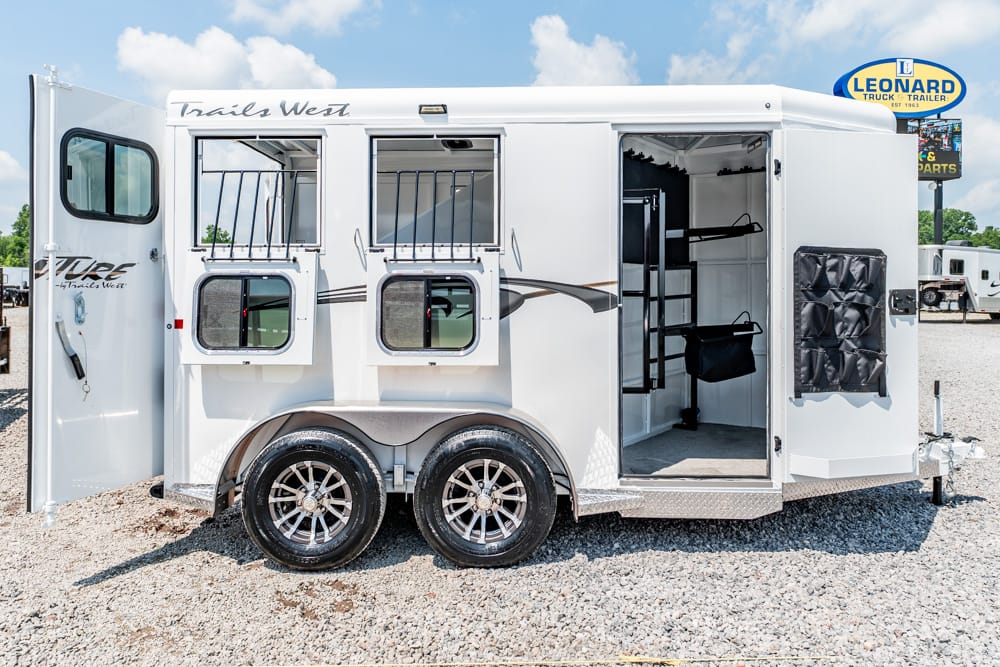 White two-axle horse trailer on a gravel lot with multiple open storage compartments and visible tack area inside camera view.