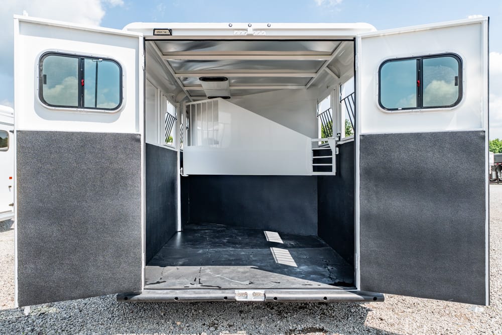 Open horse trailer interior with a white divider, side windows, and dark lower walls; rear doors are wide open.