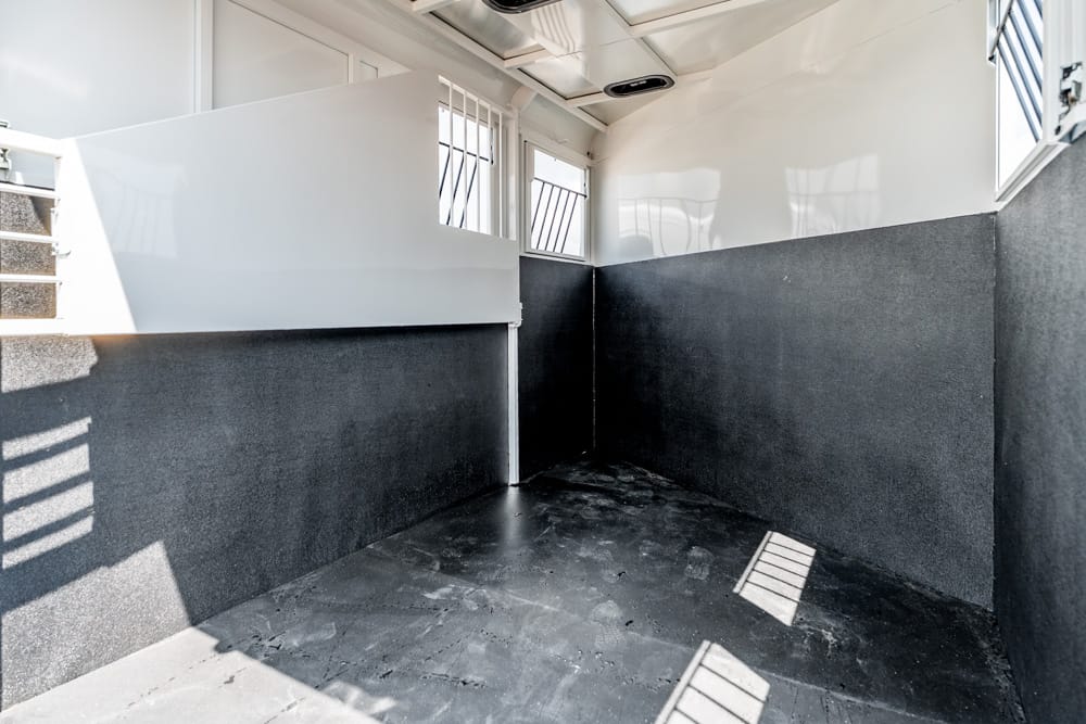 Interior of an empty horse trailer stall: dark grey lower walls, white divider, small barred windows, and a rubber mat floor with sunlight stripes