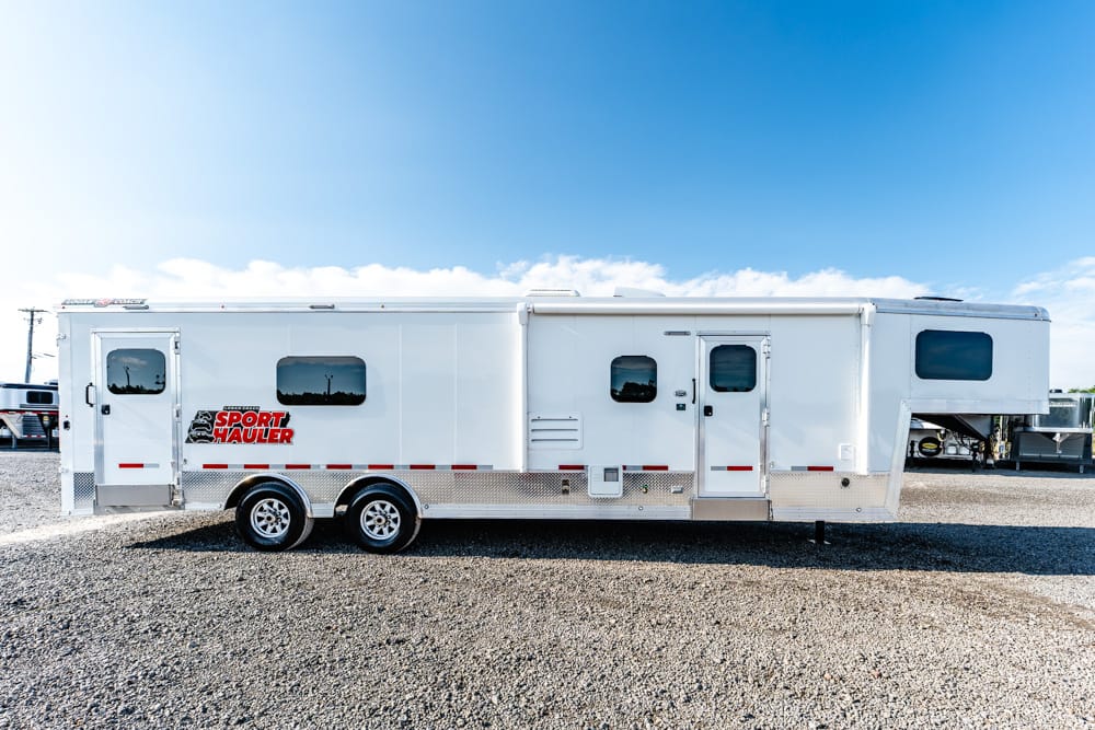 White gooseneck toy hauler trailer with two axles, side windows, and a red Sport Hauler logo parked on a gravel lot under a blue sky.