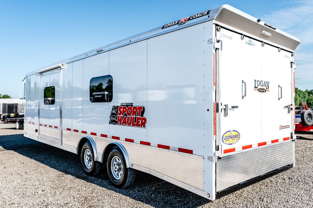 White sport hauler trailer with red 'Sport Hauler' logo on the side, dual chrome wheels, diamond-plate front, parked on gravel road