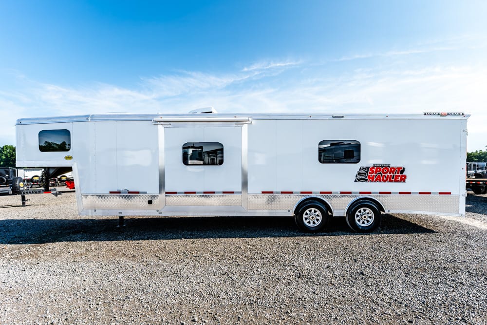 White sport hauler trailer with dual axles parked on gravel under a blue sky.