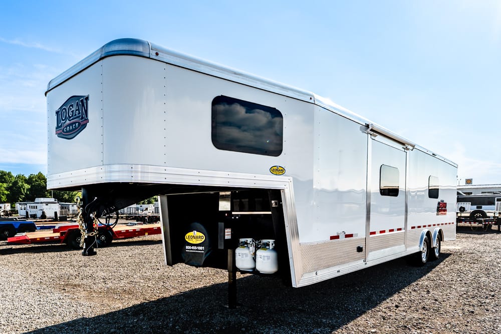 White gooseneck horse trailer with Logan Coach logo parked on a gravel lot; front storage compartment is open showing propane tanks and equipment.