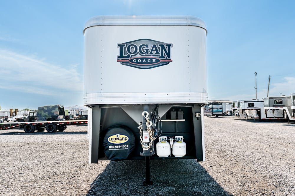 Front of a white Logan Coach gooseneck trailer with spare tire cover and propane tanks at the hitch area on a gravel lot.
