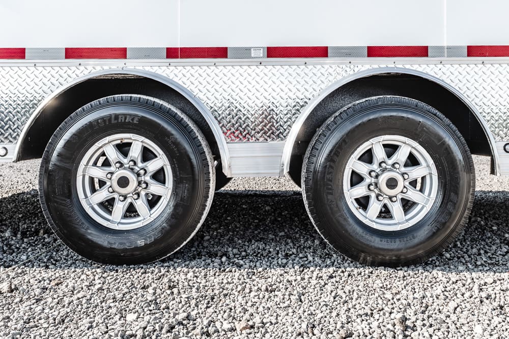 Dual-axle trailer with two black tires and silver rims on a gravel surface, side-by-side under a diamond-plate fender area.