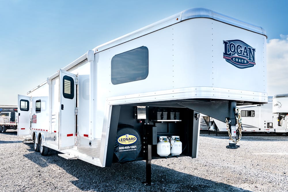 White Logan Coach fifth-wheel trailer parked on gravel, showing the front hitch and propane tanks.