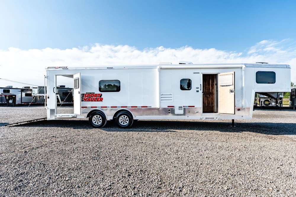White sport hauler trailer with side and rear doors open, parked on a gravel lot under a blue sky (dual-axle RV-style trailer).
