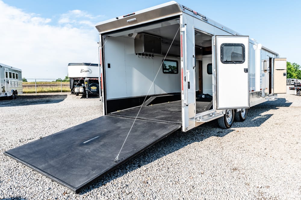 Aluminum multi-axle trailer with its rear ramp lowered and open interior on a gravel lot.