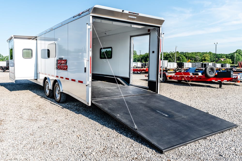 White sport hauler trailer with a wide ramp lowered for loading, parked on a gravel lot with other trailers in the background.