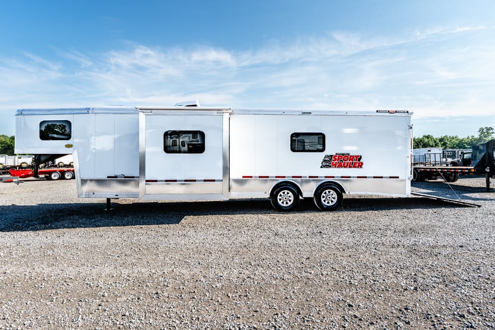 White sport hauler trailer with two axles parked on a gravel lot under a blue sky; features small windows and a side entry door near the middle with a logo reading 'SPORT HAULER' on the side.