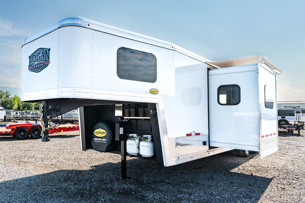 White Logan Coach gooseneck travel trailer with a side slide-out; storage bay open showing propane tanks and equipment on gravel lot