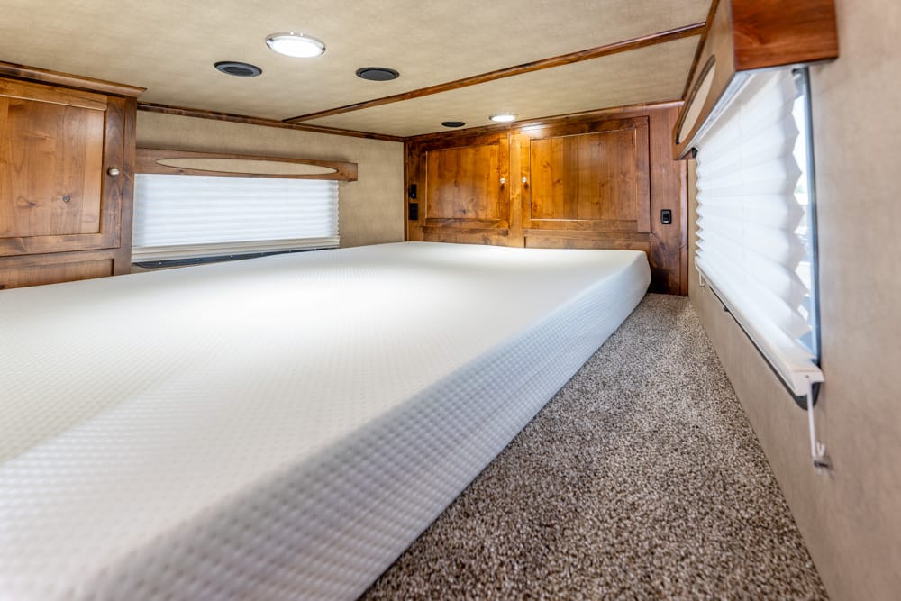 Wood-panelled RV bedroom with a long white mattress, overhead cabinets, and windows with blinds.