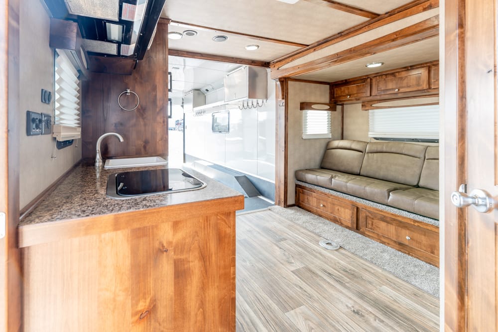Interior of a compact RV: wood kitchen counter with sink and stove on the left, beige sofa on the right with windows above.