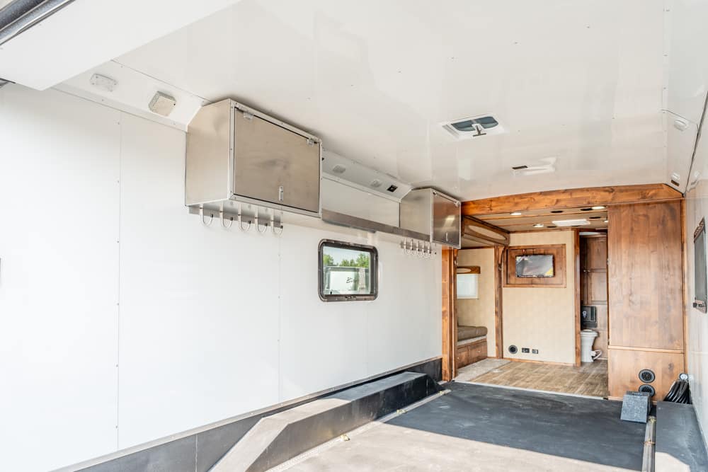 Open trailer interior with white walls, stainless steel overhead cabinets and hooks along the wall, leading to a wood-paneled living area at the rear.