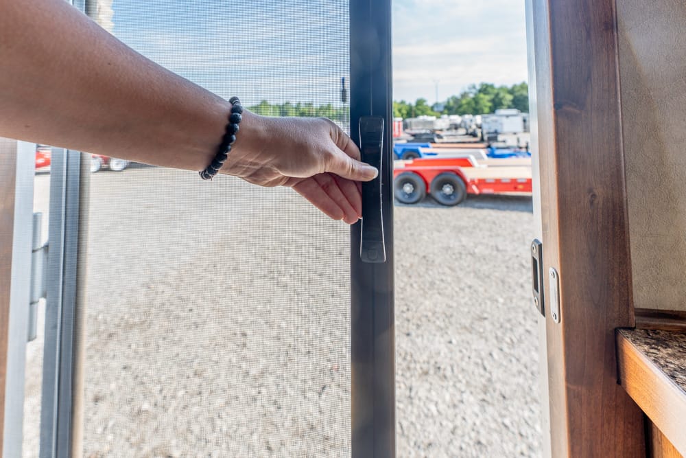 Close-up of a hand with a beaded bracelet turning a door handle to open a glass door, exposing a gravel lot with trailers outside.