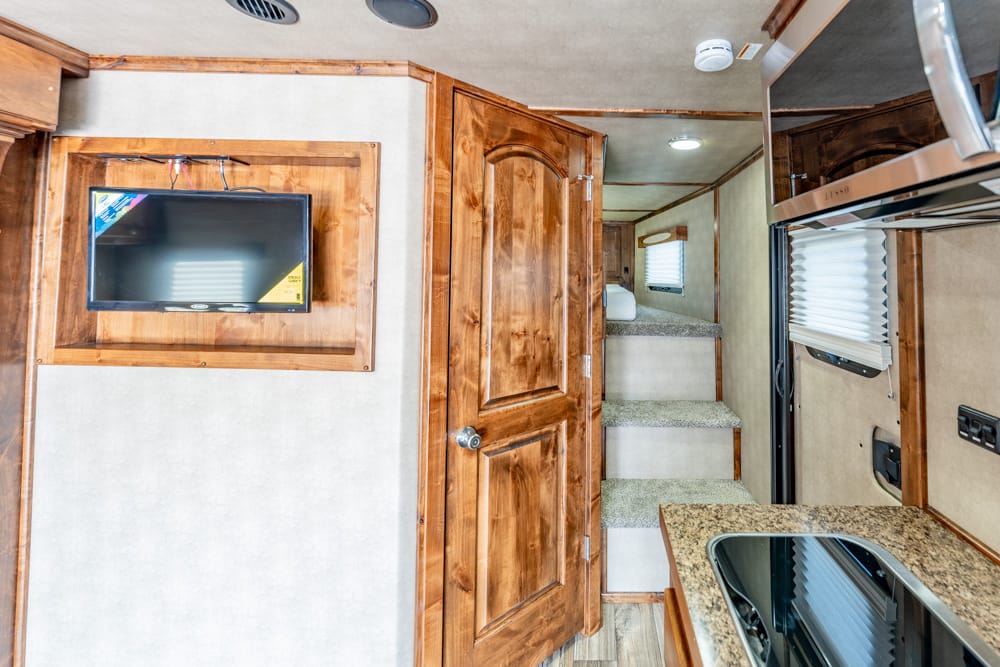 Interior of a wood-toned RV with a wall-mounted TV, a closed wooden door, and carpeted stairs ahead to a higher level.