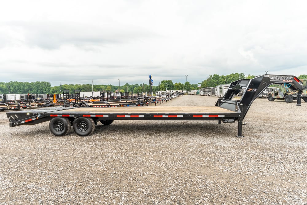 Long flatbed trailer with a gooseneck hitch sits on a gravel lot among rows of trailers and equipment.