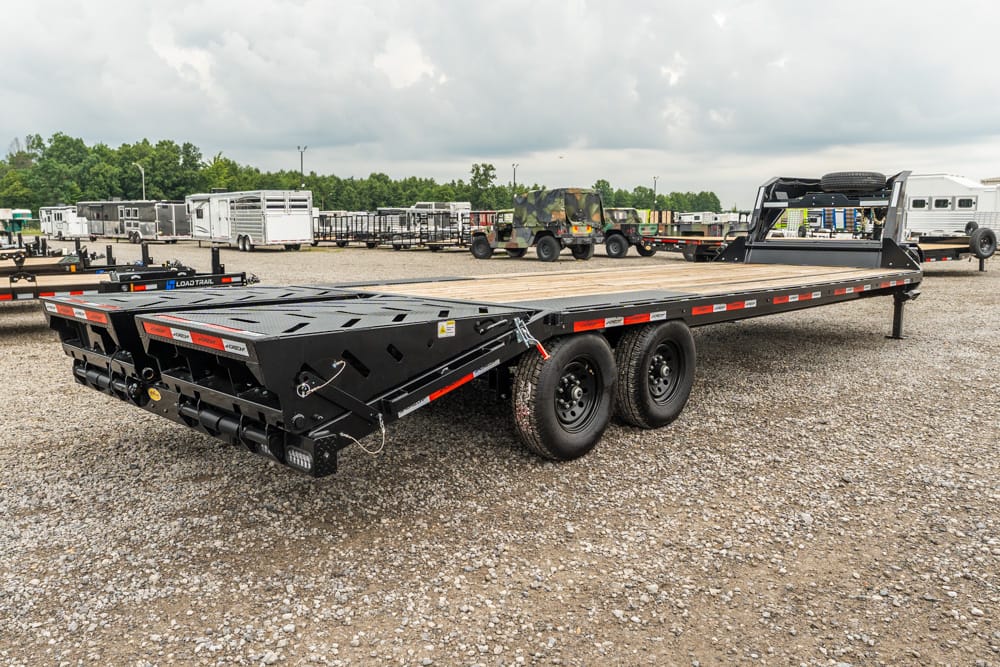 Black heavy-duty flatbed trailer with dual rear axles and gooseneck hitch, parked on a gravel lot with other trailers in the background.