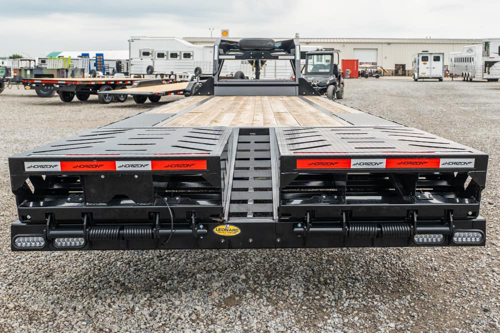 Flatbed trailer with dual ramps and rear deck, branded Horizon, parked on a gravel lot with equipment and trailers in the background.