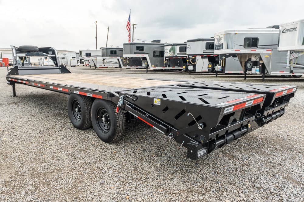 Black multi-axle tilt equipment trailer on a gravel lot with RVs in the background; long flatbed with wooden deck and ramp sections at the far end.