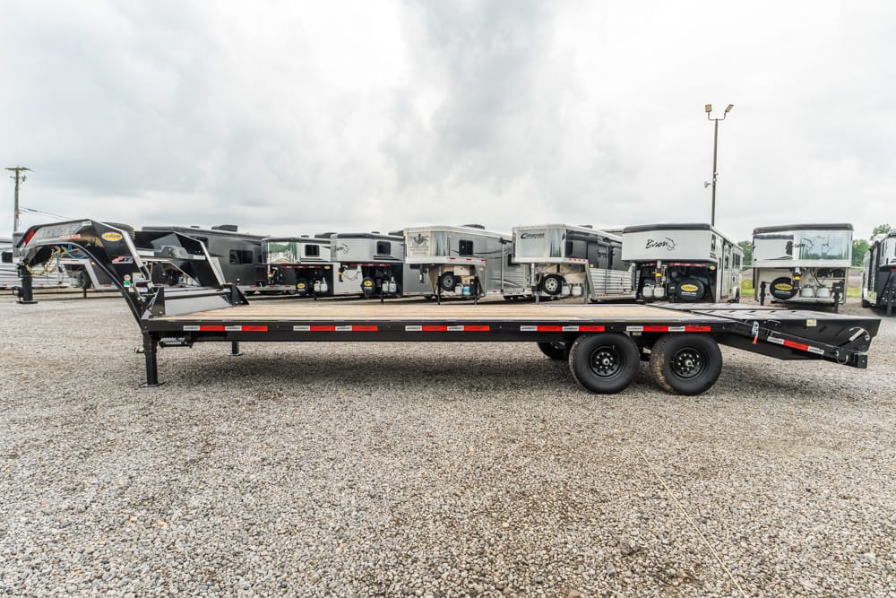 Gooseneck heavy-duty flatbed trailer on gravel lot, with a row of travel trailers parked behind.