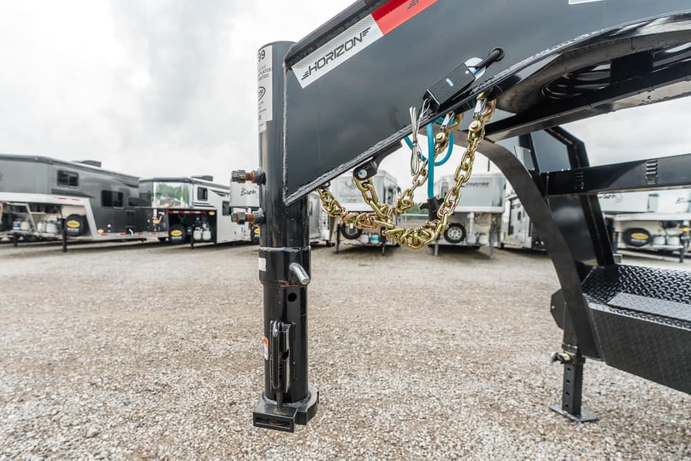 Close-up of a gooseneck trailer hitch with safety chains attached in a gravel RV lot with campers in the background.