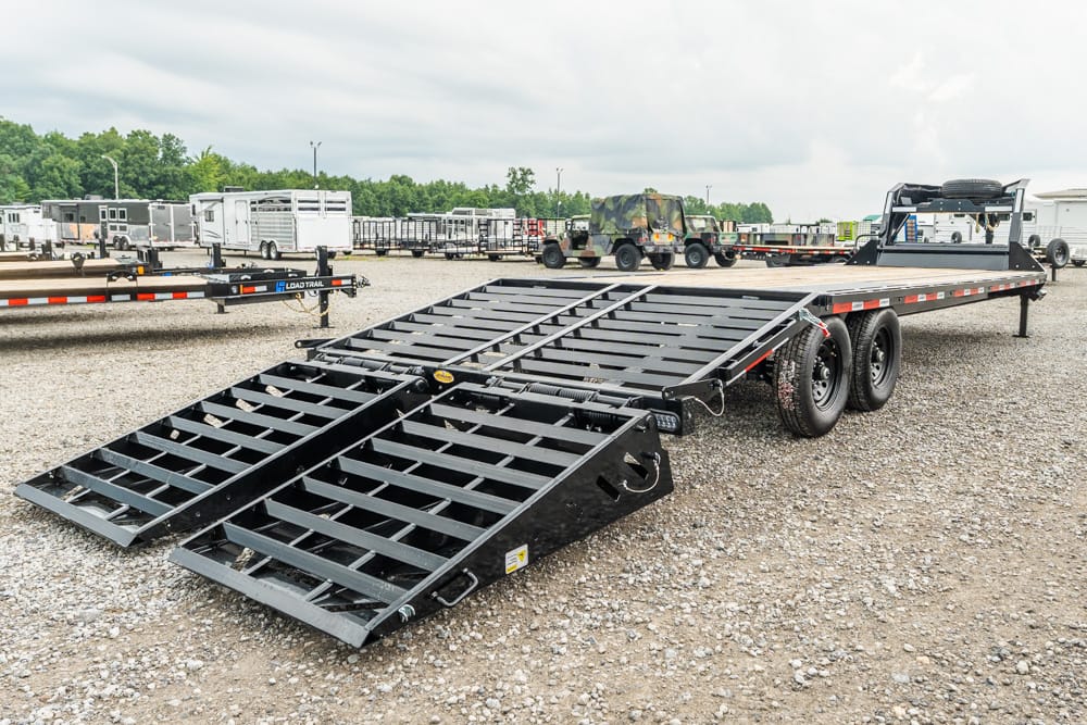 Flatbed car trailer with two heavy ramps lowered for loading, parked on a gravel lot with other trailers in the background.