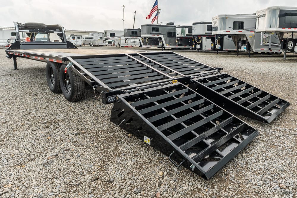 Black equipment trailer with fold-down ramps deployed on a gravel lot, with other trailers visible in the background.