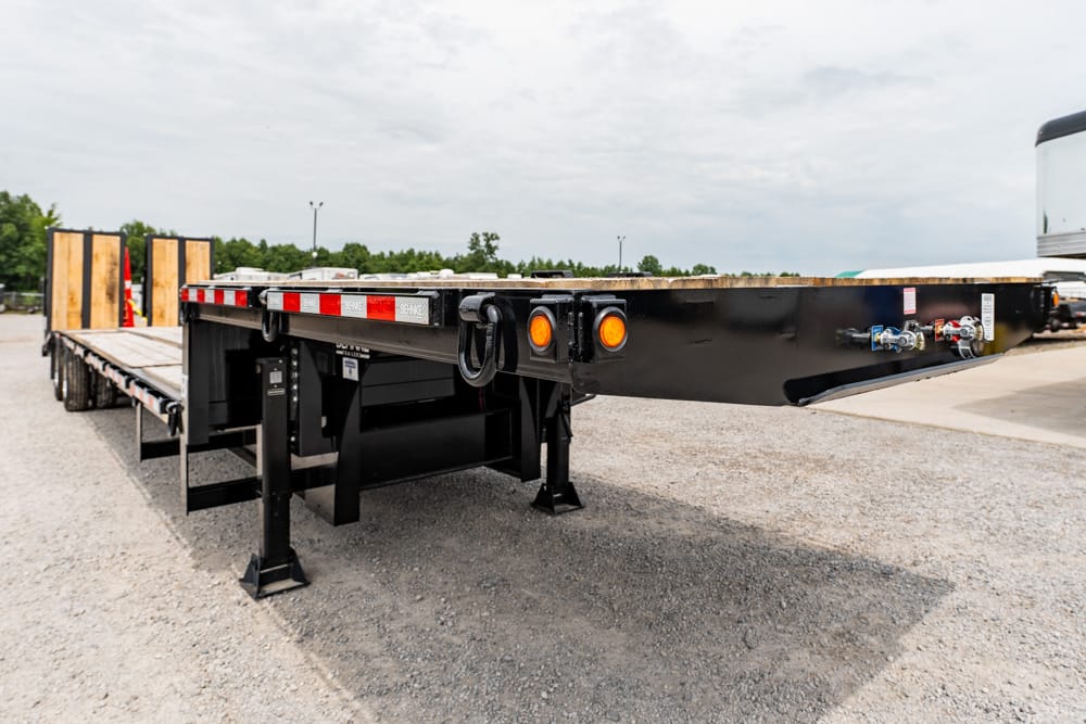 Low-angle view of a black flatbed trailer with a wooden deck, metal frame, and orange marker lights in a gravel lot.