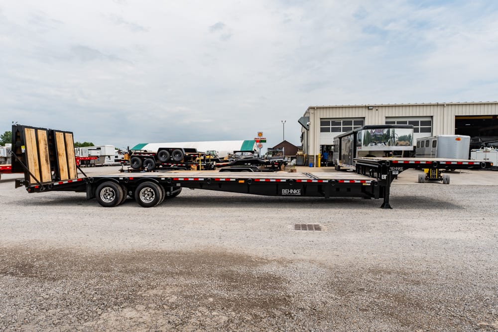 Long black flatbed trailer with a rear ramp on a gravel lot, BEHNKE branding, in an industrial yard with a workshop in the background.