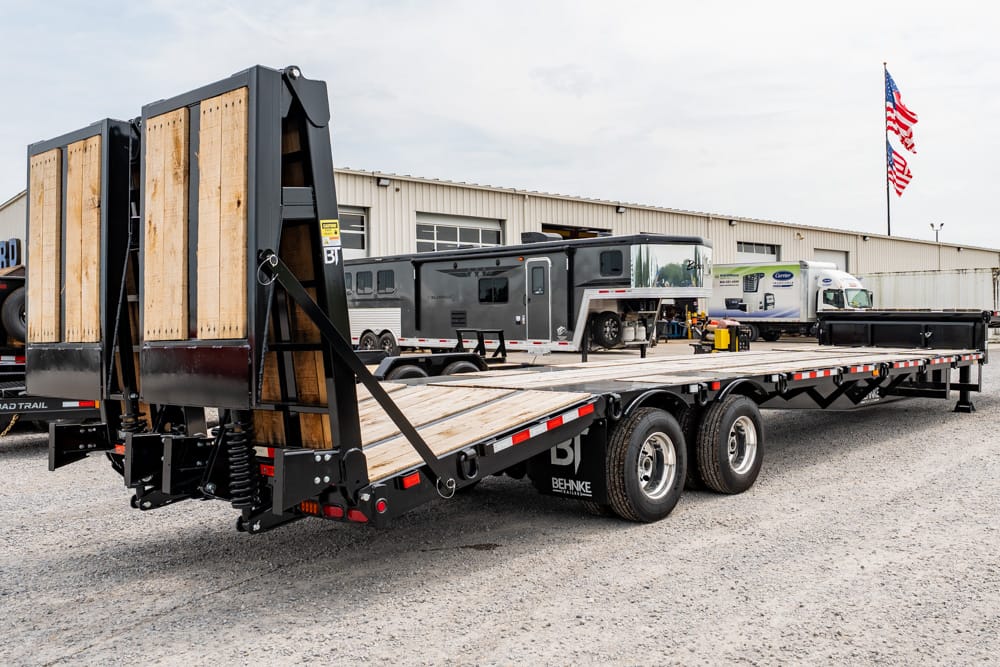 Large flatbed trailer with rear loading ramps in a truck yard, several rigs parked nearby and American flags in the background